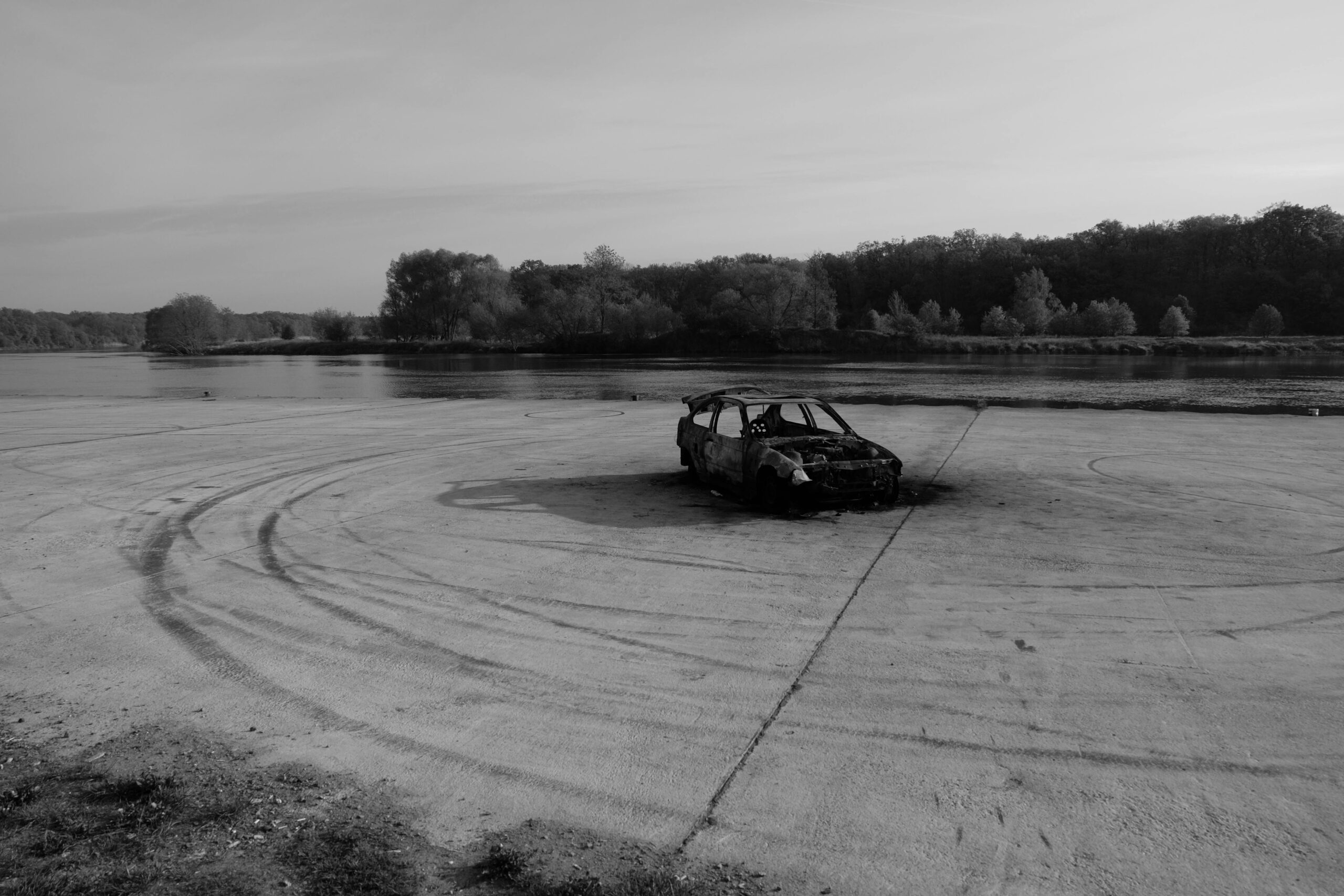 Black and white photo of a burnt car by a riverbank in Wrocław, emphasizing decay.